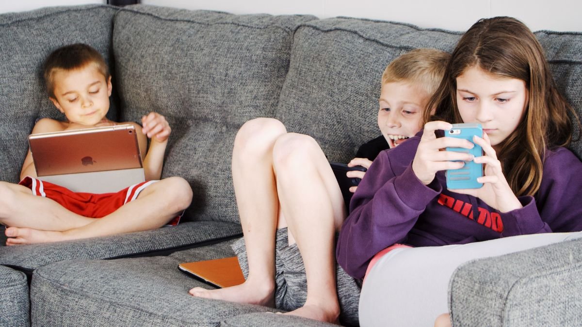 Drei Kinder sitzen auf einem Sofa und nutzen digitale Geräte, ein Tablet und ein Smartphone.
