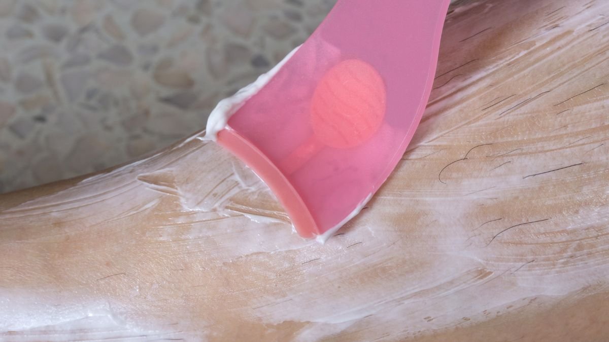 Eine Hand, die Enthaarungscreme mit einem pinken Spachtel auf die Haut aufträgt.