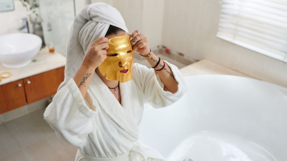 Eine Person in einem weißen Bademantel hält eine goldene Gesichtsmaske vor ihr Gesicht.