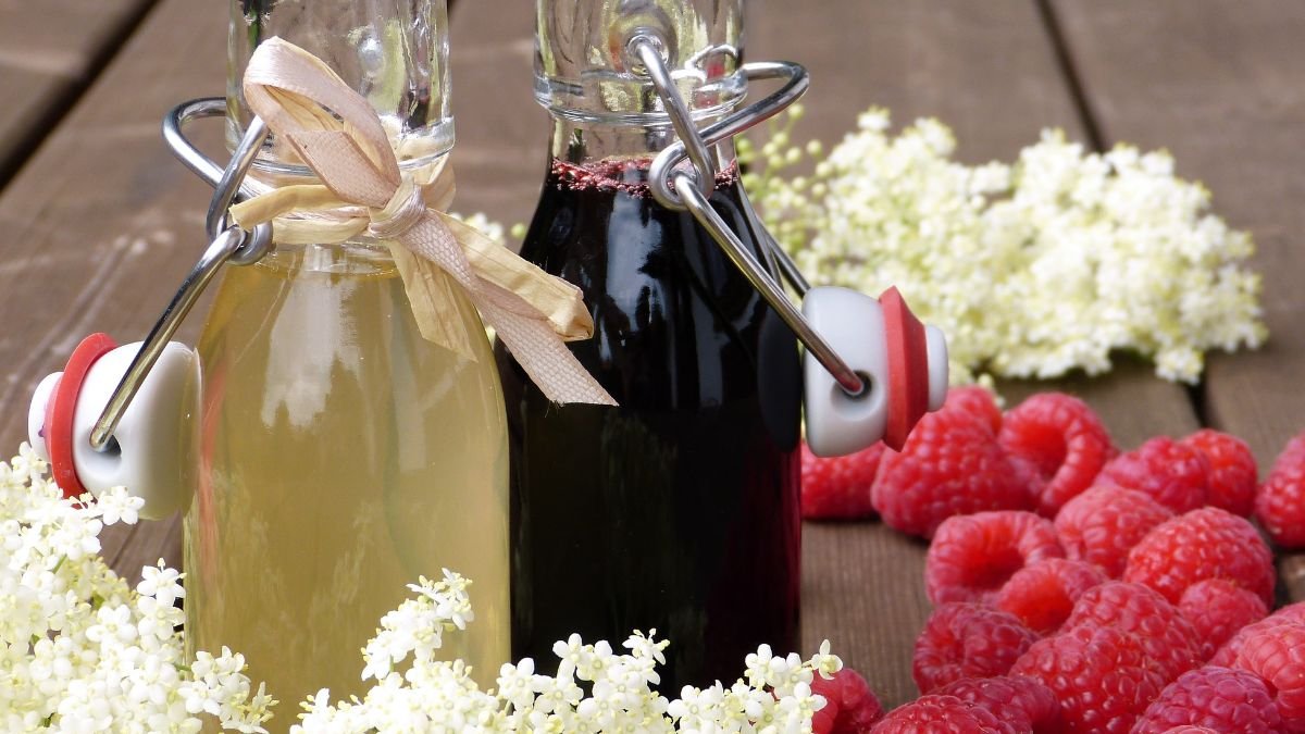 Zwei Flaschen Holundersirup, eine klare und eine dunkle, umgeben von frischen Himbeeren und Holunderblüten.