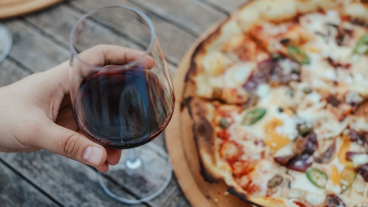 Ein Glas Rotwein wird neben einer frisch gebackenen Pizza gehalten.