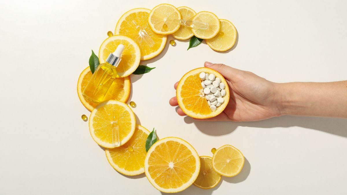 Eine Hand hält eine halbierte Orange mit Vitamin C-Tabletten, umgeben von Orangenscheiben und einem Ölfläschchen.