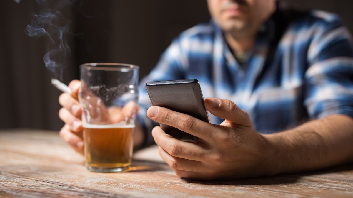 Ein Mann hält ein Bierglas in der einen Hand und ein Smartphone in der anderen, während Rauch aufsteigt.