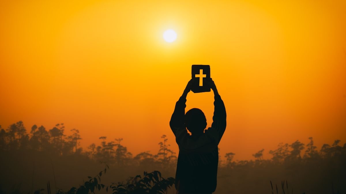 Eine Person hält eine Bibel mit einem Kreuz vor einem Sonnenuntergang.