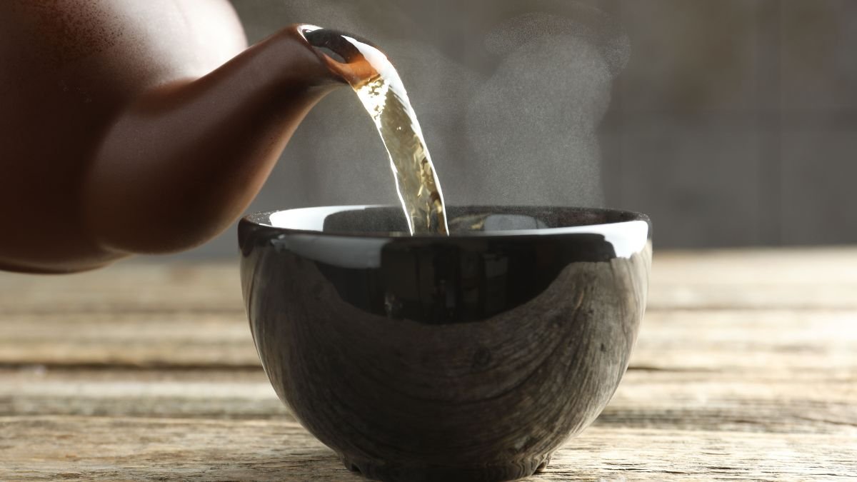 Eine Teekanne gießt dampfenden Gänsefingerkraut-Tee in eine schwarze Tasse.