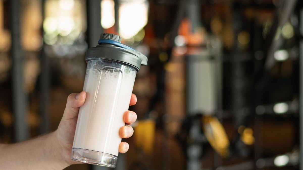 Ein Hand hält einen Shaker mit schaumigem, hellen Proteinshake im Fitnessstudio.