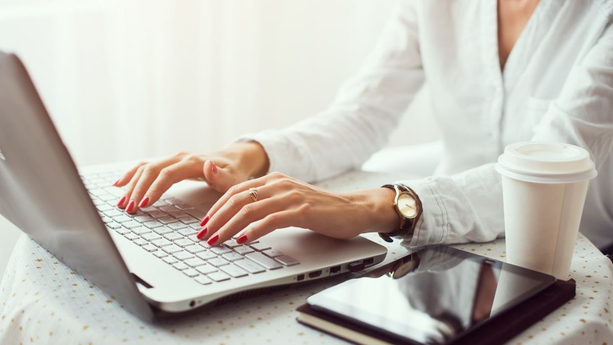 Eine Person tippt konzentriert auf einem Laptop, umgeben von Kaffee und einem Tablet.