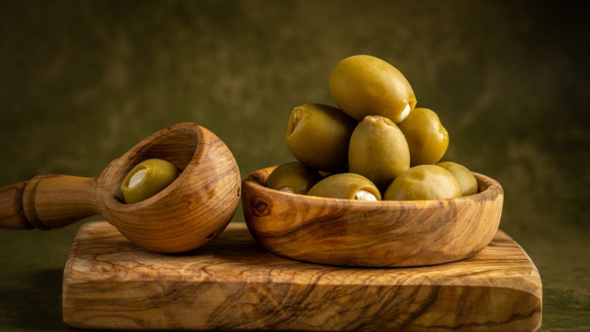 Eine Schale mit grünen Oliven auf einem Holzbrett, daneben ein Holzlöffel.