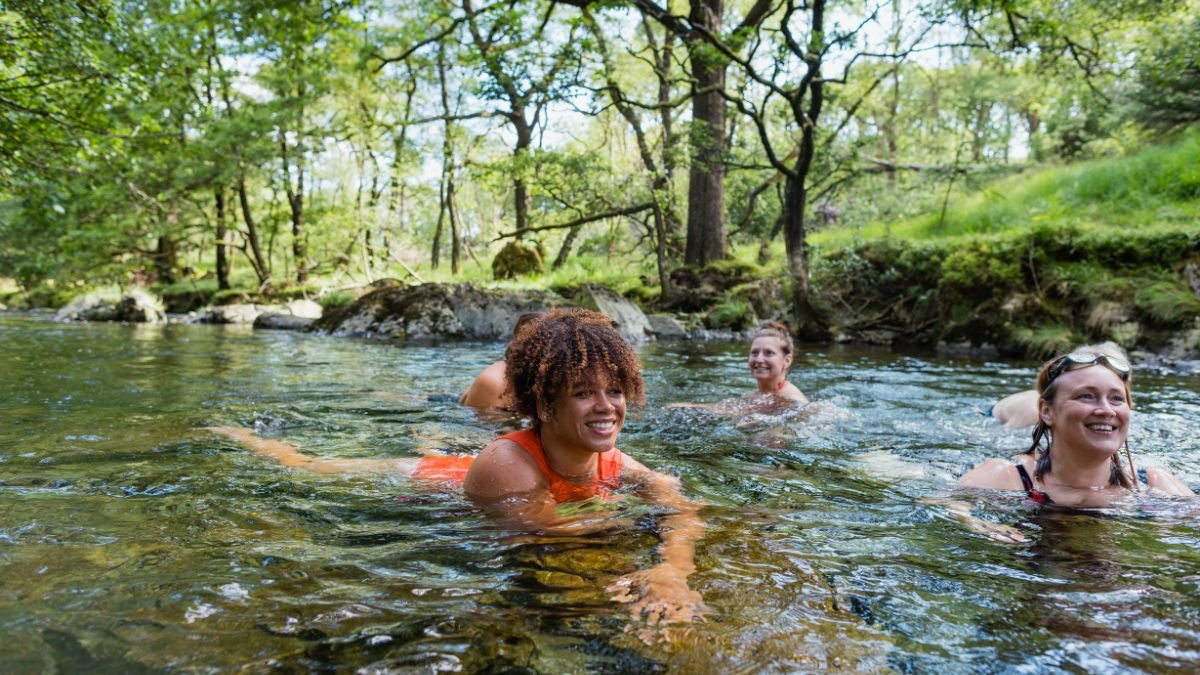 Drei Frauen schwimmen fröhlich in einem klaren, kühlen Gewässer umgeben von Bäumen.