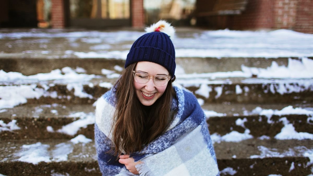 Eine lächelnde Frau in einer blauen Mütze und Decke sitzt auf schneebedeckten Stufen.