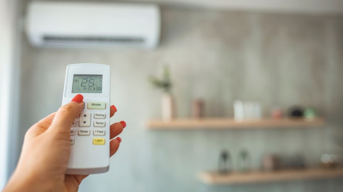 Eine Hand hält eine Fernbedienung mit einer Klimaanlage im Hintergrund.