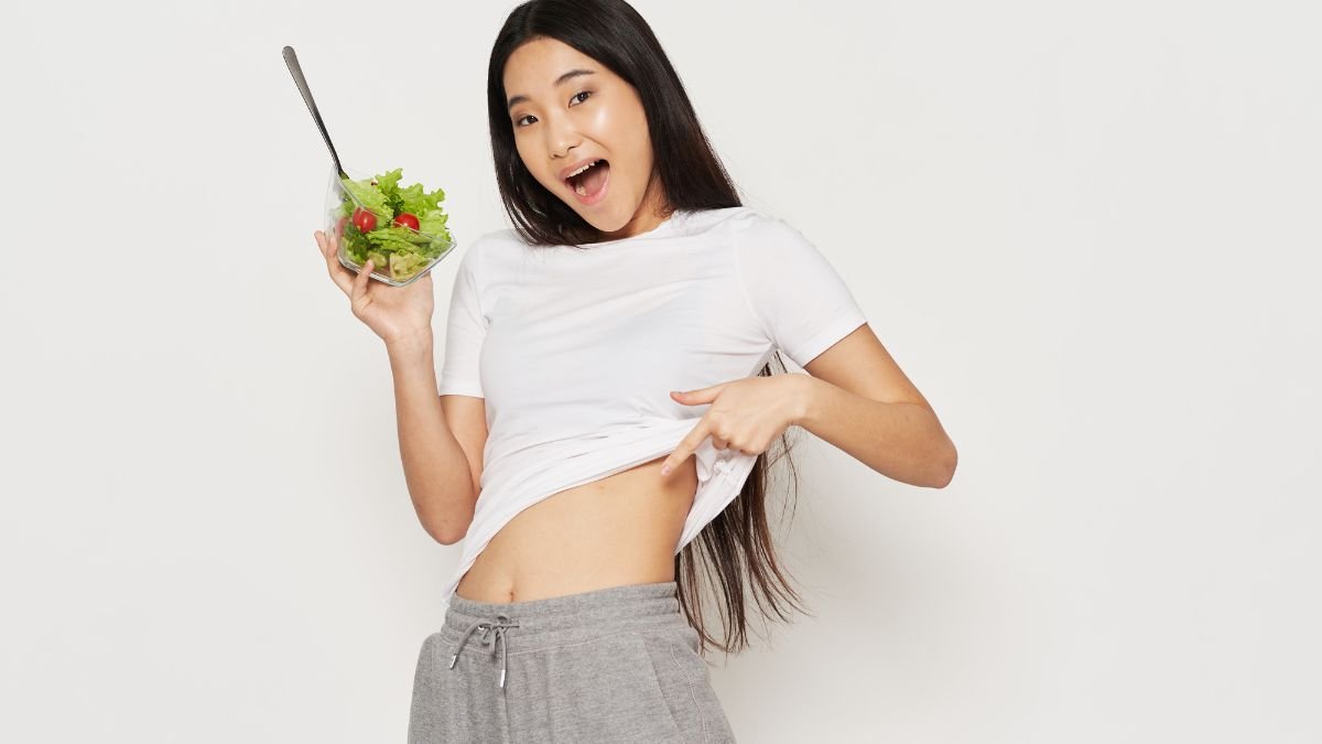 Eine junge Frau zeigt stolz ihren flachen Bauch und hält eine Schüssel mit Salat.