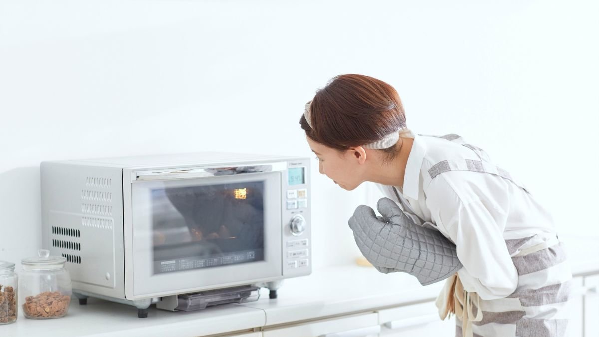 Eine Frau schaut neugierig in eine Mikrowelle, während sie Ofenhandschuhe trägt.