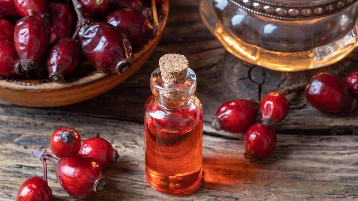 Eine Flasche Rosenöl steht neben frischen Hagebutten auf einem Holztisch.
