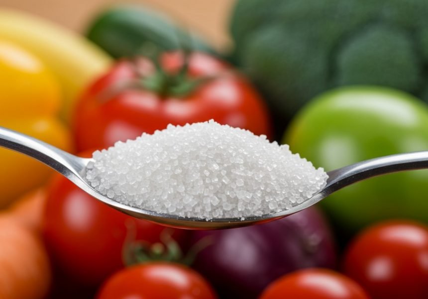 Ein Löffel mit Zucker vor einer bunten Auswahl an frischem Gemüse.