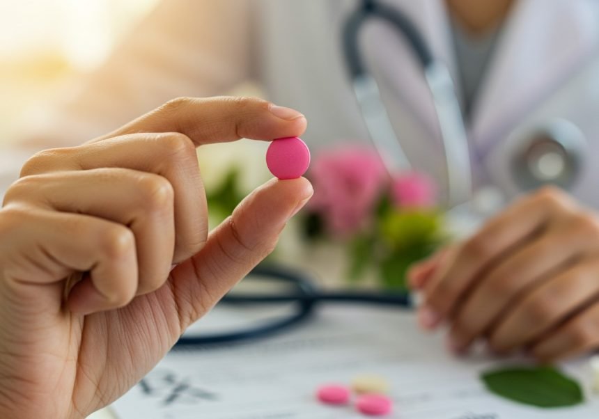 Eine Hand hält eine pinke Tablette, während ein Arzt im Hintergrund zu sehen ist.